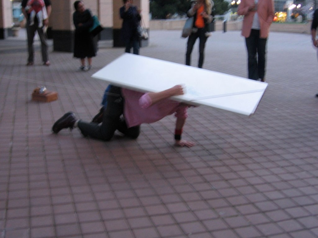 Blurry photo of a man crawling while under a white board in a square while onlookers watch