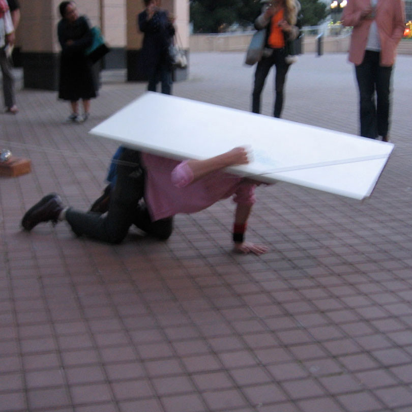 blurred photo of a man crawling with a white board on his back, in a plaza as onlookers watch.